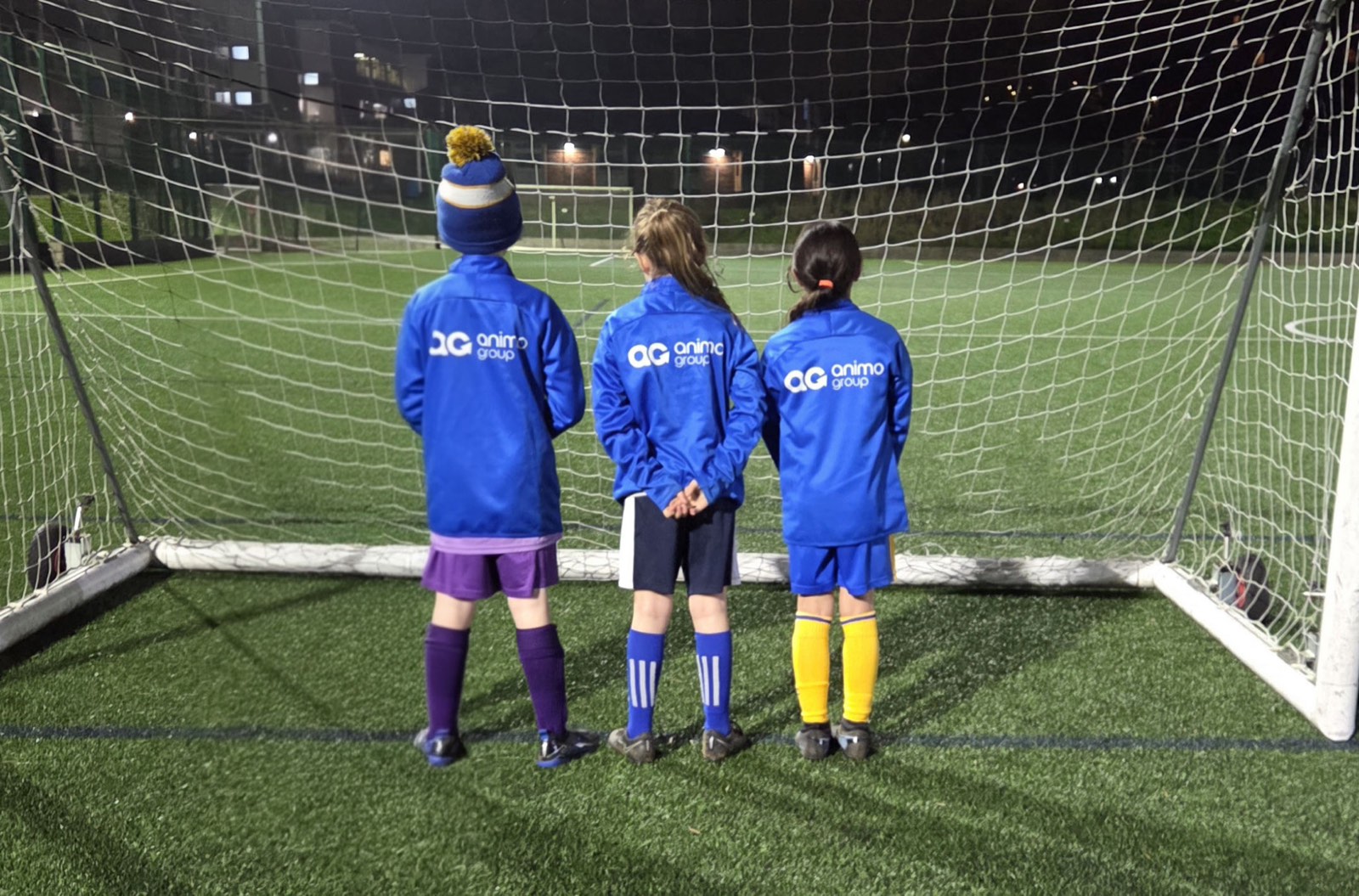 Three young players in Animo Group branded jackets standing in front of the goal at an evening training session